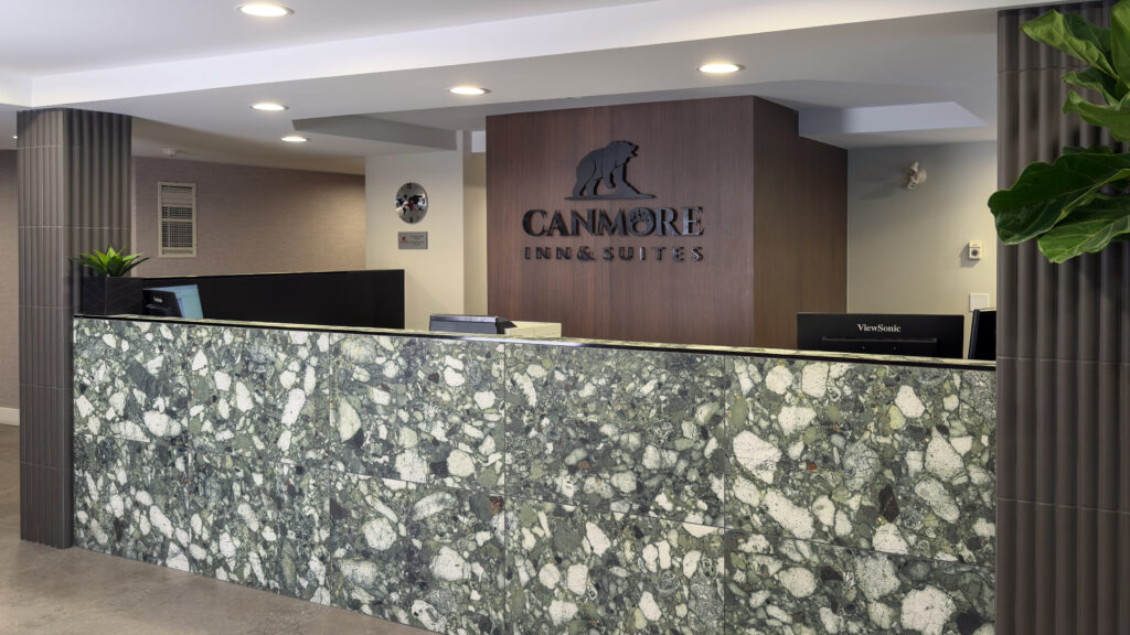 Renovated front desk at Canmore Inn & Suites featuring a modern green stone reception counter, dark wood paneling with the hotel logo, soft ambient lighting, and contemporary decor.